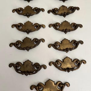May include: A collection of nine matching brass drawer pulls arranged in rows. Each pull features a central decorative backplate with a curved, ornate silhouette and a swinging bail handle attached to two circular mounting points on the sides.