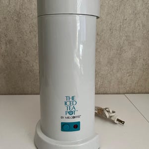 May include: A white cylindrical iced tea maker with a removable top lid. The front features the teal text THE ICED TEA POT BY MR. COFFEE above a rectangular teal power button with a small red indicator light. A white power cord is attached.