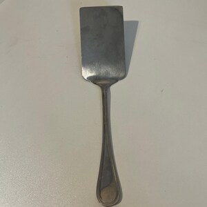 May include: A stainless steel spatula with a rectangular head and a long, slender handle. The handle has a teardrop-shaped end. The spatula is designed for cooking and food preparation.