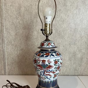May include: A decorative ceramic table lamp with a white base, adorned with red, blue, and gold floral and bird motifs. The lamp features a brass neck and a bulb, resting on a black wooden base. The lamp's design is reminiscent of Asian art.