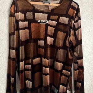 May include: A long-sleeve shirt in a brown and tan block pattern with a velvet-like texture. The chest features the white text VERSACE INTENSIVE. The shirt has a round neckline and side slits at the hem, hanging on a wooden clothes hanger.