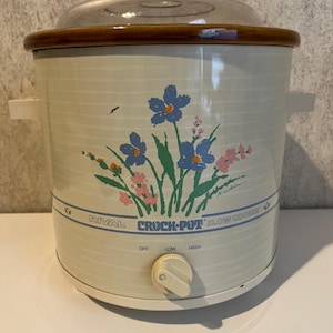 May include: A cream-colored Rival Crock-Pot slow cooker with a clear glass lid and a brown rim. The exterior features a floral graphic with blue, pink, and green flowers, and a control knob with settings for off, low, and high heat levels.