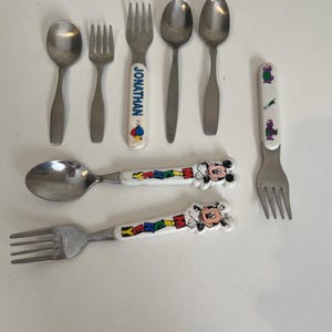 May include: A collection of children's silverware, including spoons and forks. Some utensils have plain metal handles, while others feature colorful designs with the name "JONATHAN", a cartoon character, and Mickey Mouse.