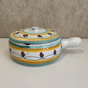May include: A ceramic soup bowl with a lid, featuring a white base with a floral design and a yellow and green striped pattern. The bowl has a white handle and a small lid with a knob.