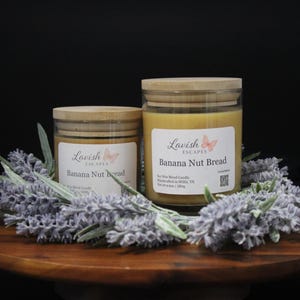 May include: Two glass jars of Banana Nut Bread scented soy wax candles with light wood lids. Each label features the Lavish Escapes logo with a butterfly graphic. The candles are surrounded by artificial lavender sprigs on a round wooden base.