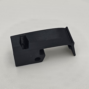 May include: A black, 3D-printed component with a rectangular base and a curved top. It has a circular hole and a notched section. The object is likely a part of a mechanism. The item is set against a white background.