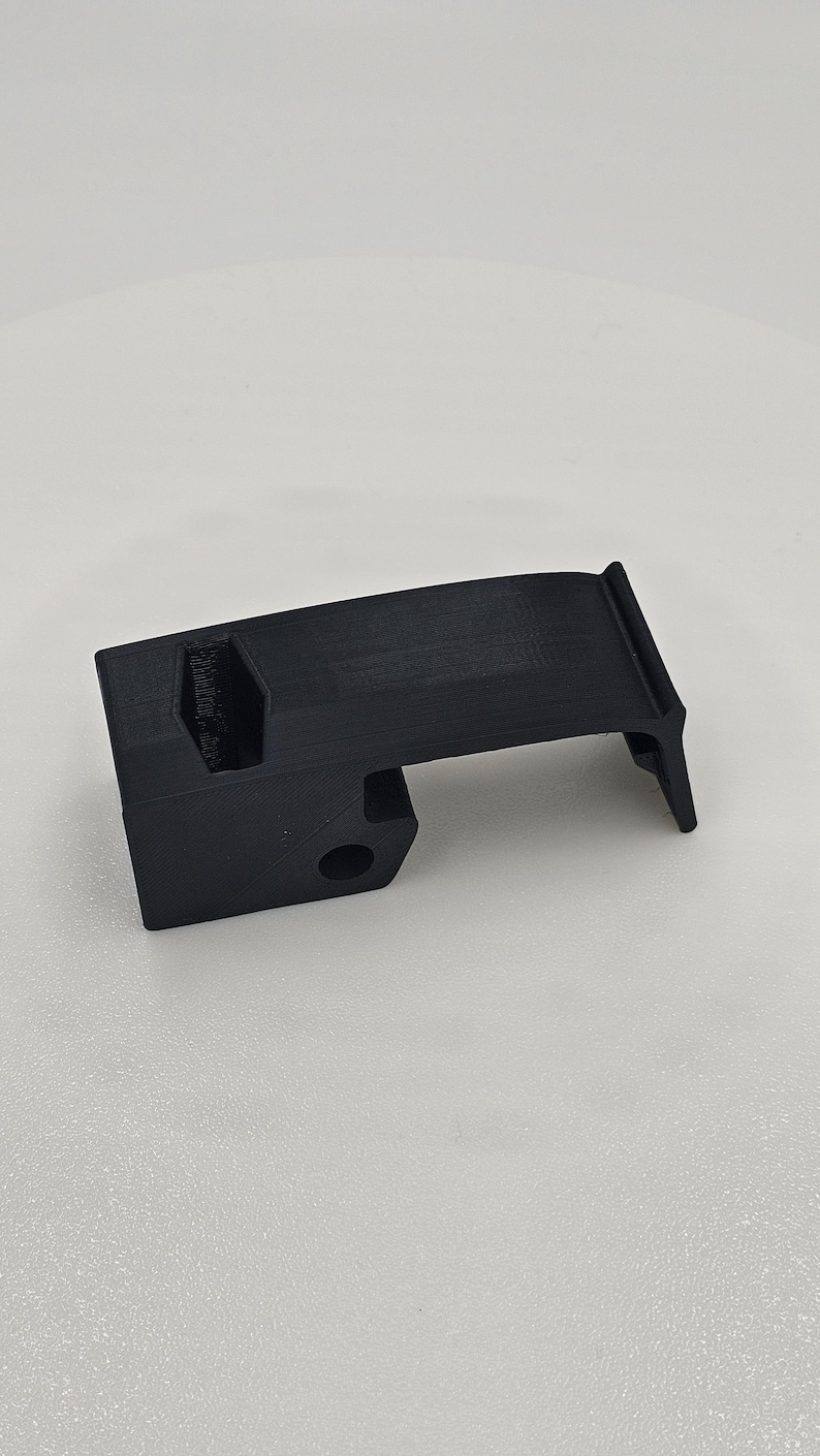 May include: A black, 3D-printed component with a rectangular base and a curved top. It has a circular hole and a notched section. The object is likely a part of a mechanism. The item is set against a white background.