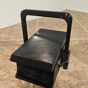 Peut inclure: Une boîte noire rectangulaire avec une poignée. La boîte a une surface texturée et une poignée qui se courbe sur le dessus. La boîte est posée sur un sol carrelé.