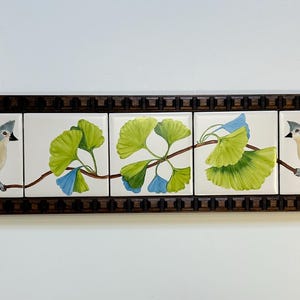 May include: A rectangular wall art piece featuring five square ceramic tiles set in a dark brown wooden frame. The tiles display a continuous hand-painted scene of two grey and yellow tufted titmouse birds perched on a branch with green ginkgo leaves.