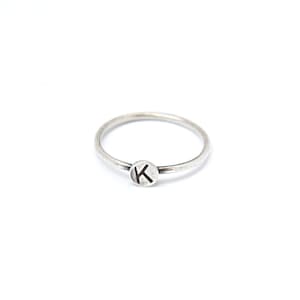 May include: A thin silver-toned ring featuring a small, circular flat charm attached to the band. The charm is engraved with the uppercase letter K in a dark, bold font. The metal has a slightly oxidized, matte finish throughout.