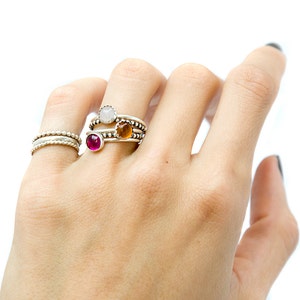 May include: A hand wearing three silver rings with different colored gemstones. The rings are stacked on the index finger. The gemstones are white, orange, and pink.
