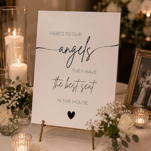 Könnte beinhalten: Ein rechteckiges weißes Schild mit schwarzem Text, der lautet: HERE'S TO OUR angels THEY HAVE the best seat IN THE HOUSE, mit einem kleinen schwarzen Herzsymbol am unteren Rand. Das Schild steht auf einer dünnen goldenen Metallstaffelei.