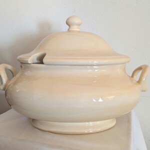 May include: Cream-coloured ceramic soup tureen with a lid and two handles. The tureen has a rounded base and a lid with a small knob. The lid has a notch for a ladle. The tureen is designed for serving soup or stew.