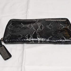 May include: A black and silver snakeskin-effect wallet with a zip closure. The rectangular wallet features a small metal detail on the top. A black tag with silver text is attached to the zip.