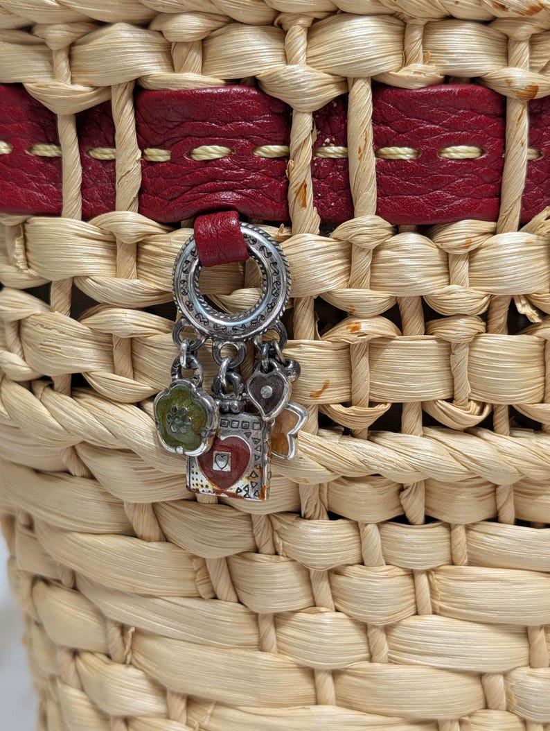 Może przedstawiać: Pleciony kosz ze słomy z poziomym czerwonym sk&oacute;rzanym paskiem z jasnymi przeszyciami. Srebrzysty metalowy charms wisi na sk&oacute;rze, prezentując kilka małych zawieszek, w tym zielony kwiat, czerwone serce i kwadratowy kształt.