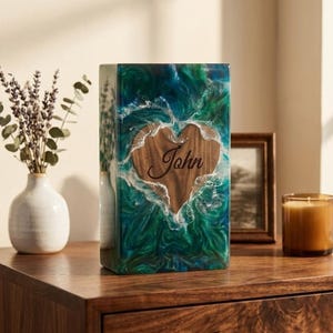May include: A rectangular, handcrafted urn with a wooden heart-shaped inlay. The heart is engraved with the name "John". The urn is encased in a vibrant, swirling mix of green, blue, and white resin, creating a unique, artistic design.