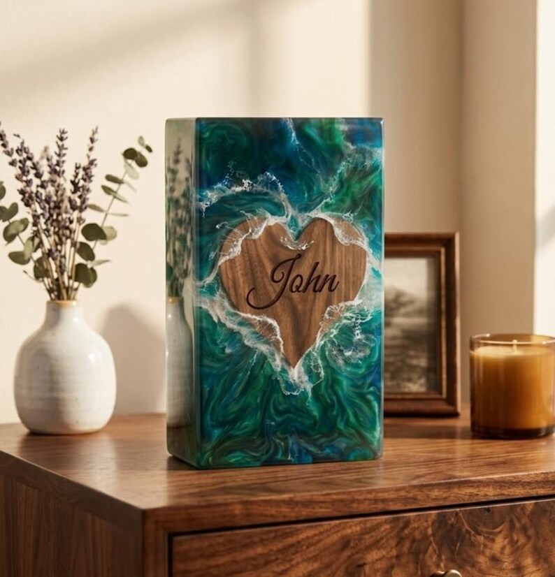 May include: A rectangular, handcrafted urn with a wooden heart-shaped inlay. The heart is engraved with the name "John". The urn is encased in a vibrant, swirling mix of green, blue, and white resin, creating a unique, artistic design.