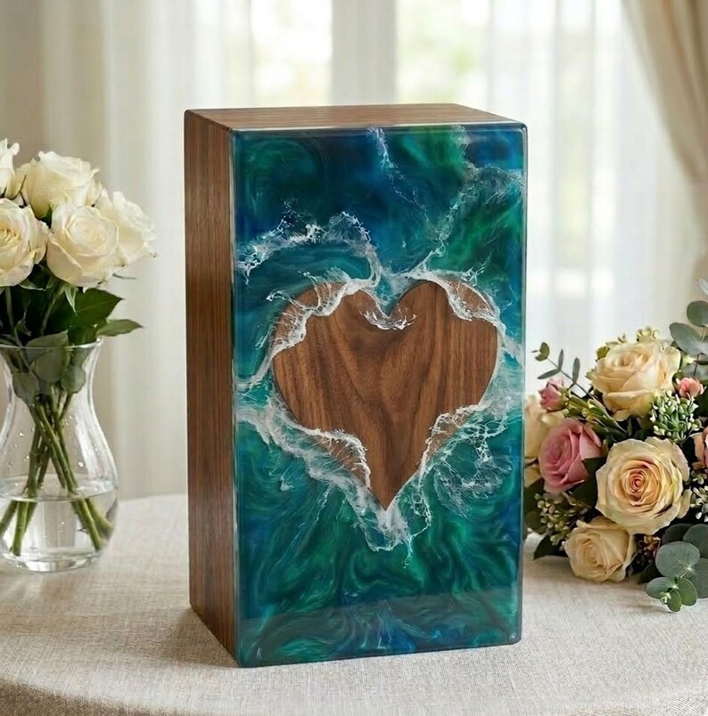 May include: A rectangular wooden box with a heart-shaped cutout. The box is encased in a blue and green resin that resembles ocean waves. The heart is made of wood. The box is sitting on a table with flowers in the background.