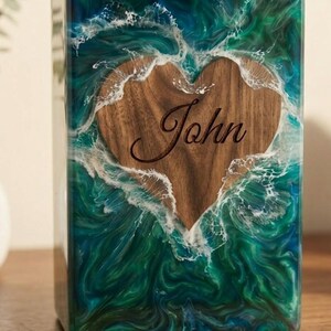 May include: A rectangular wooden box with a heart-shaped cutout. The heart is engraved with the name "John". The box is encased in a resin design with blue and green swirls, resembling ocean waves.
