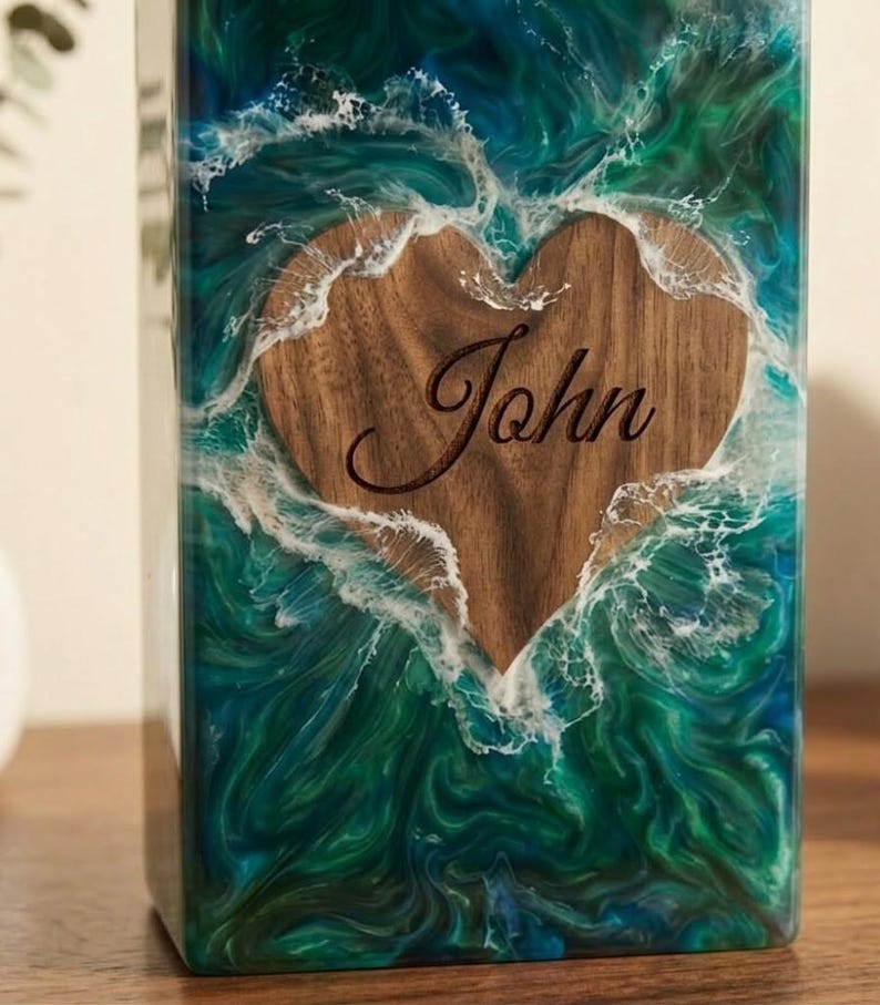May include: A rectangular wooden box with a heart-shaped cutout. The heart is engraved with the name "John". The box is encased in a resin design with blue and green swirls, resembling ocean waves.