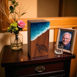 Pode incluir: Uma urna funerária retangular de madeira com acabamento em grão de madeira castanho escuro. A secção superior apresenta um design de onda oceânica em resina azul e branca, enquanto a parte inferior inclui uma gravação de uma tartaruga marinha.
