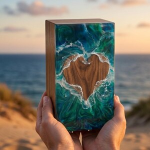 May include: A wooden box with a heart-shaped cutout, filled with blue and green resin that resembles ocean waves. The box is held up against a beach and ocean background.