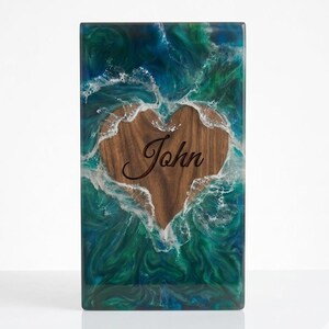 May include: Rectangular wooden box with a heart-shaped cutout. The heart is filled with wood and the name "John" is engraved. The box is surrounded by a blue and green resin design that resembles ocean waves.