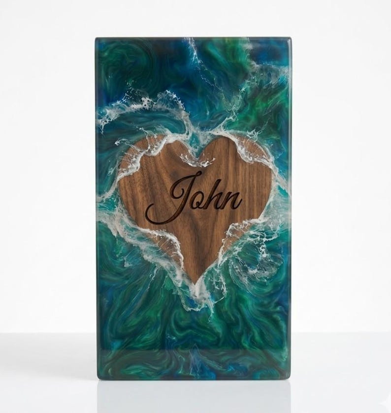May include: Rectangular wooden box with a heart-shaped cutout. The heart is filled with wood and the name "John" is engraved. The box is surrounded by a blue and green resin design that resembles ocean waves.