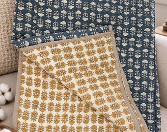 Op de afbeelding: Een omkeerbare gewatteerde plaid met een donker marineblauwe zijde met een herhalend crèmekleurig bloemenpatroon en een contrasterende crèmekleurige zijde met een herhalend mosterdgeel bloemenpatroon, afgewerkt met een effen beige rand.
