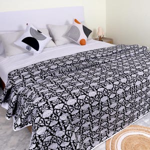 May include: A black and white quilt featuring a dense floral pattern covers a bed. Two white throw pillows with black and orange geometric arc designs are placed against plain white pillows on a white bed sheet set.
