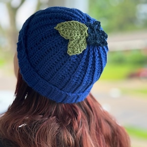May include: A handmade, blue crochet beanie with a green leaf accent. The hat is ribbed and has a folded brim. The leaf is a lighter shade of green and adds a touch of whimsy to the design. This hat is perfect for cool weather.