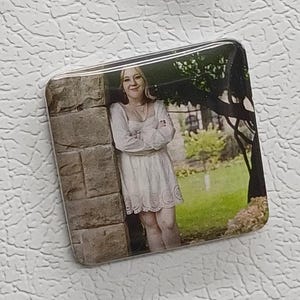 May include: A square magnet featuring a photograph of a person in a white dress leaning against a stone wall. The background includes a tree and green foliage. The magnet has a glossy finish.