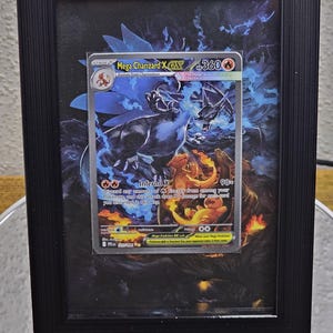 Puede incluir: Una carta Pokémon Mega Charizard X EX enmarcada. La carta presenta una ilustración de dragón azul y naranja con el texto "Inferno X". El marco negro se muestra sobre una superficie reflectante.