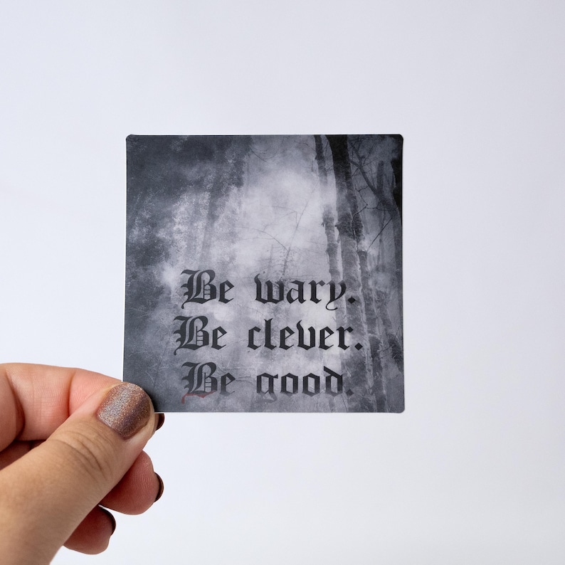 May include: Square sticker with a forest scene background and the text "Be wary. Be clever. Be good." in a gothic font. The sticker is held by a hand with painted nails.