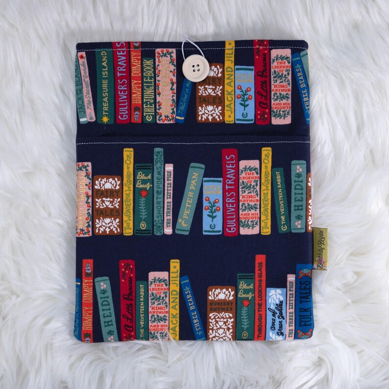 May include: A navy blue book sleeve featuring a colorful bookshelf design. The fabric is patterned with various book titles, including "Gulliver's Travels" and "Heidi." A white button and loop closure secures the sleeve, which is designed to protect books.