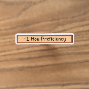 May include: A rectangular sticker with a brown border and the text "+1 Hoe Proficiency" in black. The sticker has a white border and is set against a wooden background.