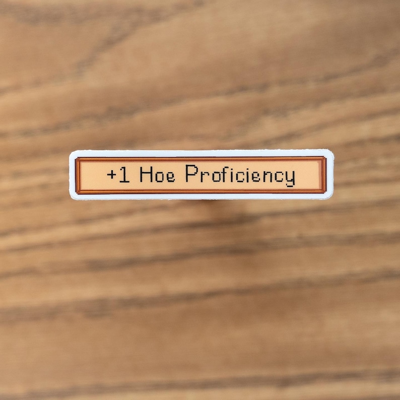 May include: A rectangular sticker with a brown border and the text "+1 Hoe Proficiency" in black. The sticker has a white border and is set against a wooden background.