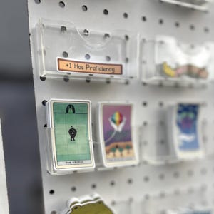 May include: Clear acrylic card holders mounted on a white pegboard. The holders contain tarot cards and stickers. One label reads "+1 Hoe Proficiency". The cards have colorful illustrations and text. The pegboard has evenly spaced holes.