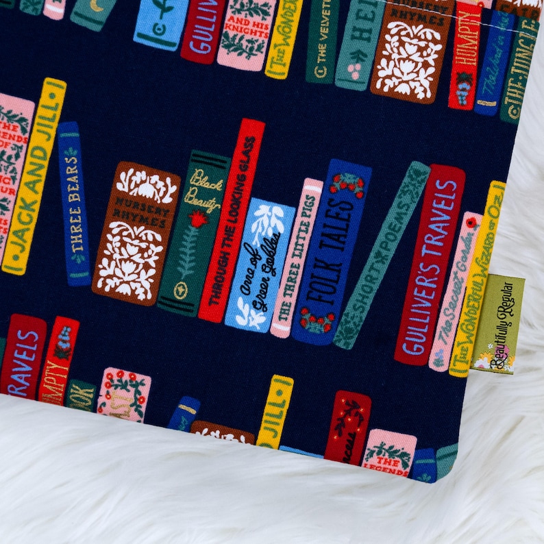 May include: A navy blue fabric featuring a colorful bookshelf design. The books have titles like "Jack and Jill," "Three Bears," and "Gulliver's Travels." The fabric has a white fluffy background and a small tag that says "Beautifully Regular."