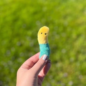 May include: A plush worm toy with a yellow head and a turquoise body. The worm has two small black dots for eyes. The toy is held in a person's hand against a blurred green background.