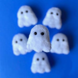 Puede incluir: Una colecci&oacute;n de seis peque&ntilde;as figuras de fantasmas de peluche blancos con ojos de bot&oacute;n negros, dispuestas sobre un fondo azul vibrante. Los fantasmas tienen una textura suave y esponjosa, y su dise&ntilde;o simple evoca un tema juguet&oacute;n y espeluznante.