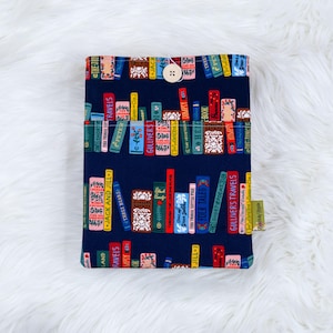 May include: A navy blue book sleeve featuring a colorful pattern of stacked books with titles like "Gulliver's Travels" and "Jack and Jill." The sleeve has a wooden button closure and a small tag.