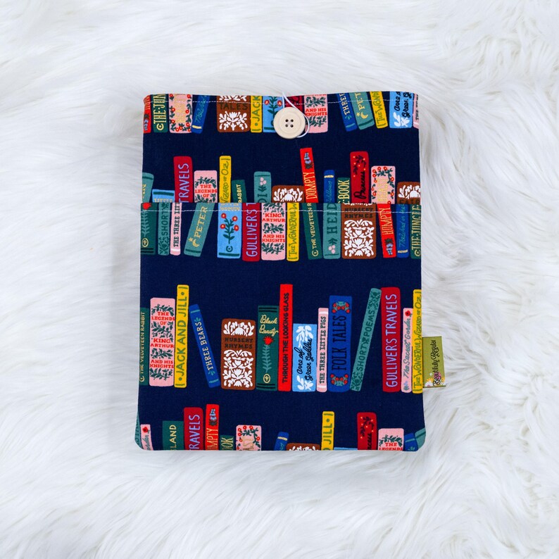May include: A navy blue book sleeve featuring a colorful pattern of stacked books with titles like "Gulliver's Travels" and "Jack and Jill." The sleeve has a wooden button closure and a small tag.