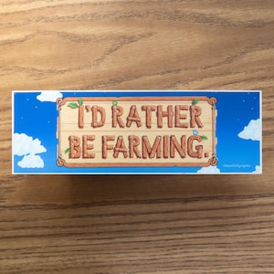 May include: A rectangular sticker with a blue sky background and white clouds. The sticker features a wooden sign with the text "I'D RATHER BE FARMING." The sign has green leaves and a small blue bird. The sticker is on a wooden surface.