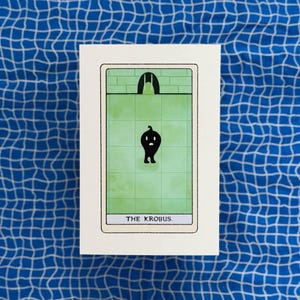 May include: A rectangular card featuring a tarot-style illustration with a light green tiled background and a dark archway at the top. A small, black, rounded creature with two legs stands in the center. The text THE KROBUS is printed at the bottom.