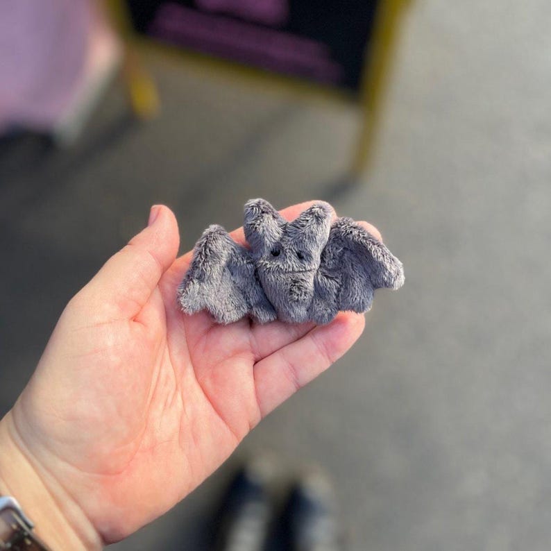 Pu&ograve; includere: Un piccolo pipistrello grigio di peluche con un volto sorridente e ali spiegate. Il pipistrello &egrave; fatto di materiale morbido e sfocato e ha un design carino e fumettistico. L'oggetto &egrave; a tema Halloween o gotico.