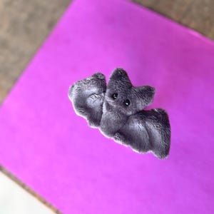 Pu&ograve; includere: Un giocattolo a forma di pipistrello grigio in peluche, con occhi neri e una superficie strutturata. Il pipistrello &egrave; posizionato su uno sfondo viola brillante, con una superficie in legno visibile nell'angolo.