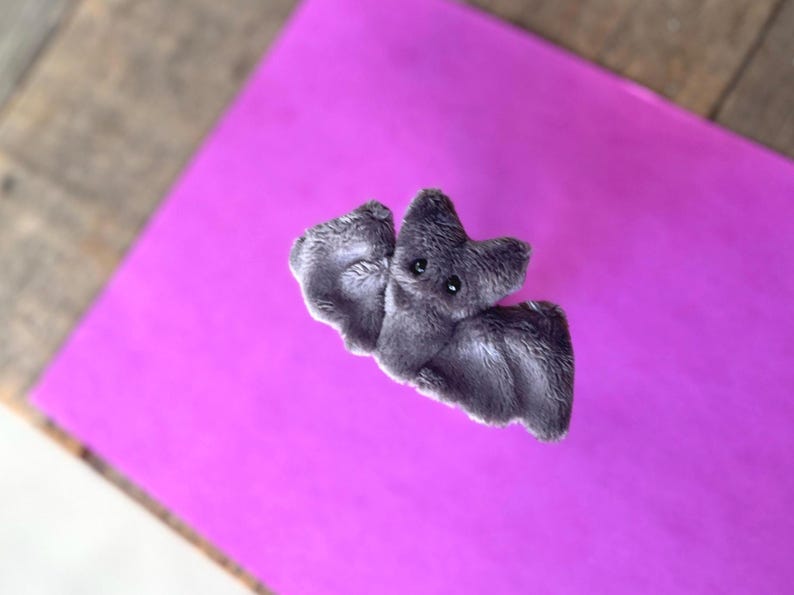 Pu&ograve; includere: Un giocattolo a forma di pipistrello grigio in peluche, con occhi neri e una superficie strutturata. Il pipistrello &egrave; posizionato su uno sfondo viola brillante, con una superficie in legno visibile nell'angolo.