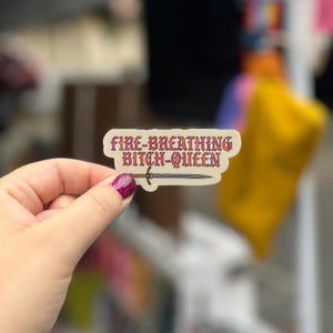 Op de afbeelding: Een sticker met de tekst "FIRE-BREATHING BITCH-QUEEN" in rode letters, met een zwaardillustratie. De sticker wordt vastgehouden in een hand met donkerroze nagellak.