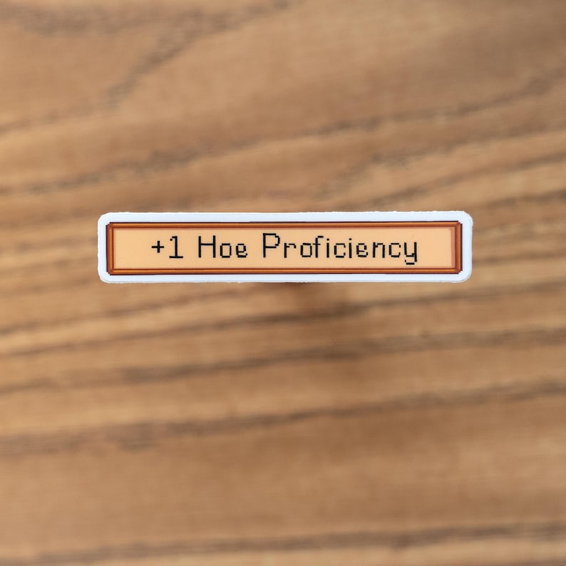 May include: A rectangular sticker with a white border and a tan center. The text "+1 Hoe Proficiency" is displayed in black pixelated font. The sticker is set against a wooden background.